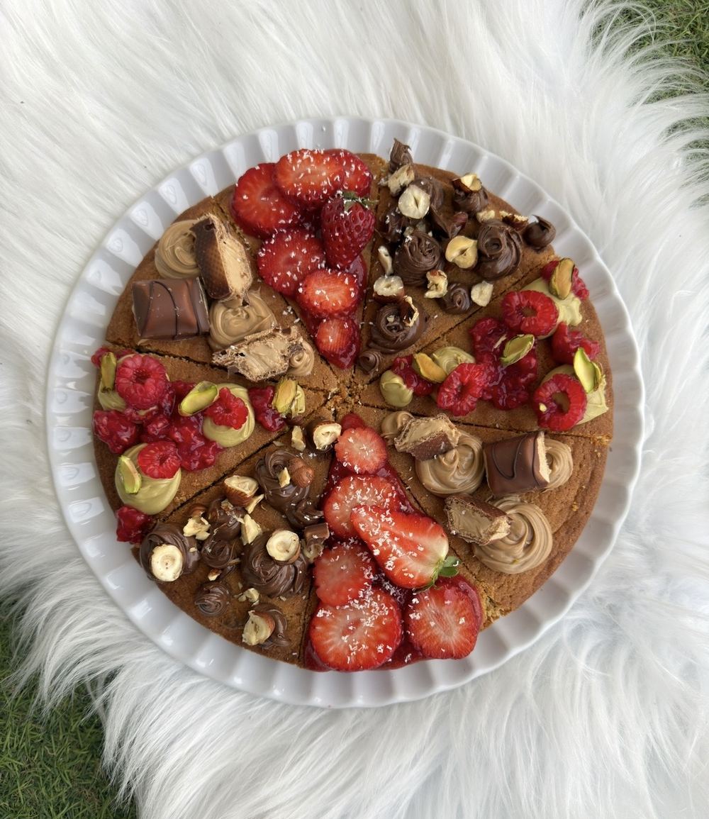 Cookie géant à partager découpé en parts, garni de fraises, framboises, crème pistache, pâte à tartiner et noisettes