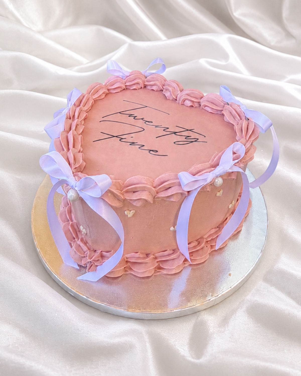 Layer cake en forme de cœur rose poudré, bordure de crème pochée, nœuds de satin lilas et inscription « Twenty Five »
